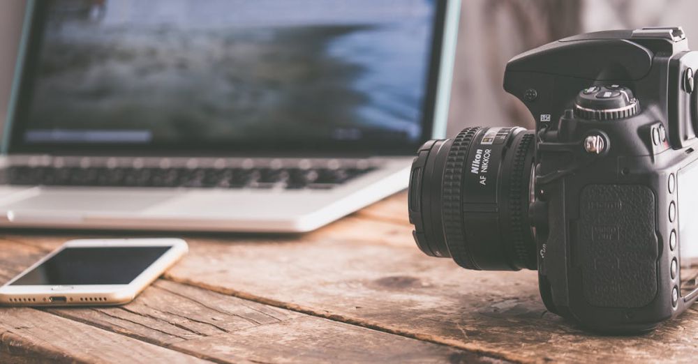 Digital Camera - Close-up of a DSLR camera, laptop, and smartphone on a wooden table in a modern workspace.