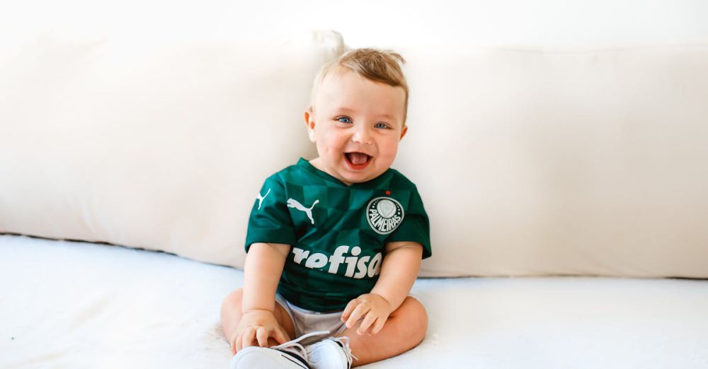 Baby Brands - Cute baby smiling while sitting on a sofa wearing a green sports jersey indoors.