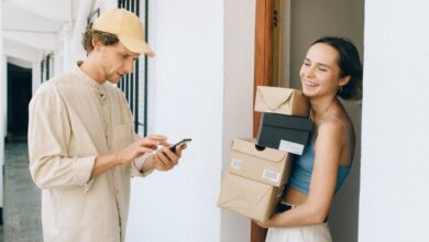 Subscription Boxes - A cheerful woman receives a delivery from a person in a hallway, using a smartphone for online payment.