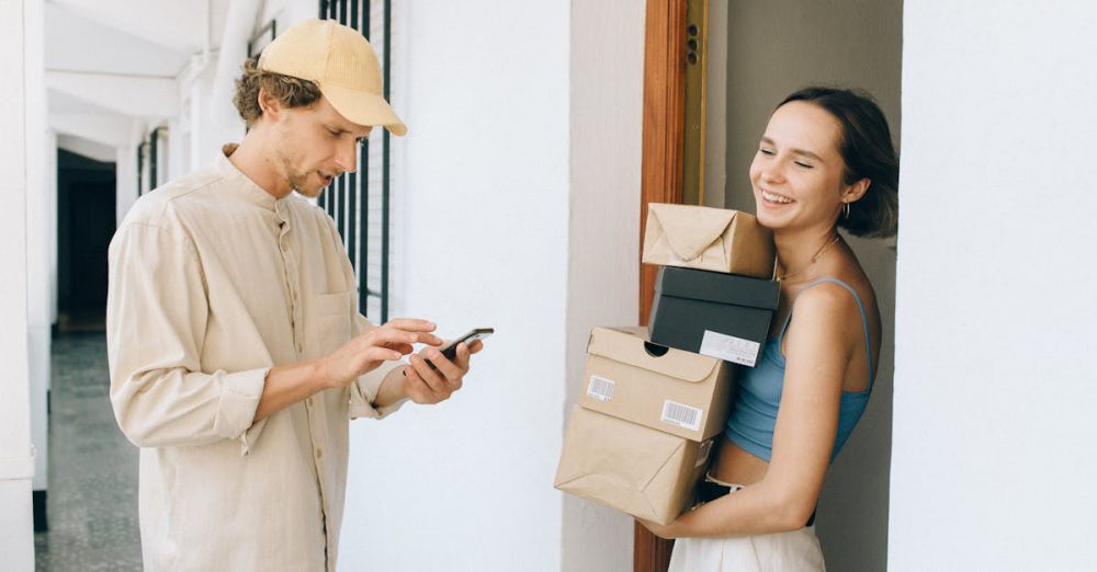 Subscription Boxes - A cheerful woman receives a delivery from a person in a hallway, using a smartphone for online payment.