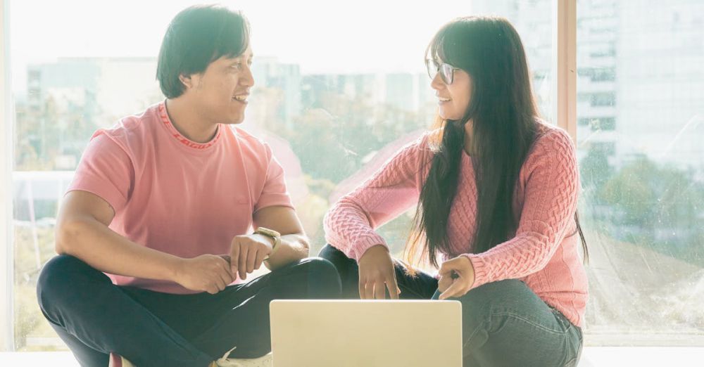 Laptop For Students - Two young adults engaged in discussion while working on a laptop near a large window.