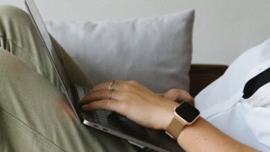 Smart Home Gadgets - Side view of crop unrecognizable female freelancer in casual clothes and smartwatch lying on comfortable sofa and working remotely on laptop