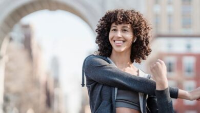 Bluetooth Earbuds - Happy ethnic sportswoman in activewear stretching arms and looking away while training on street with historic arch on blurred background