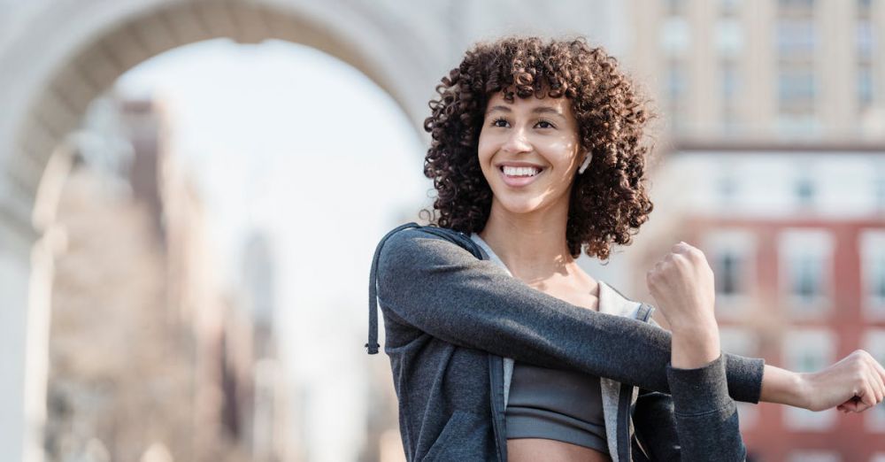 Bluetooth Earbuds - Happy ethnic sportswoman in activewear stretching arms and looking away while training on street with historic arch on blurred background