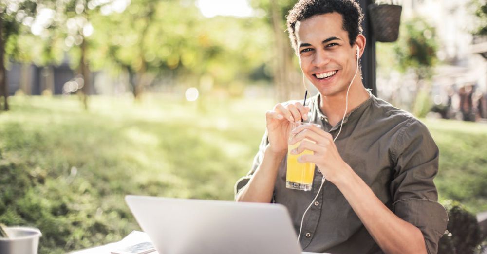 Smart Headphones - Cheerful guy with laptop and earphones sitting in park while drinking juice and smiling at camera