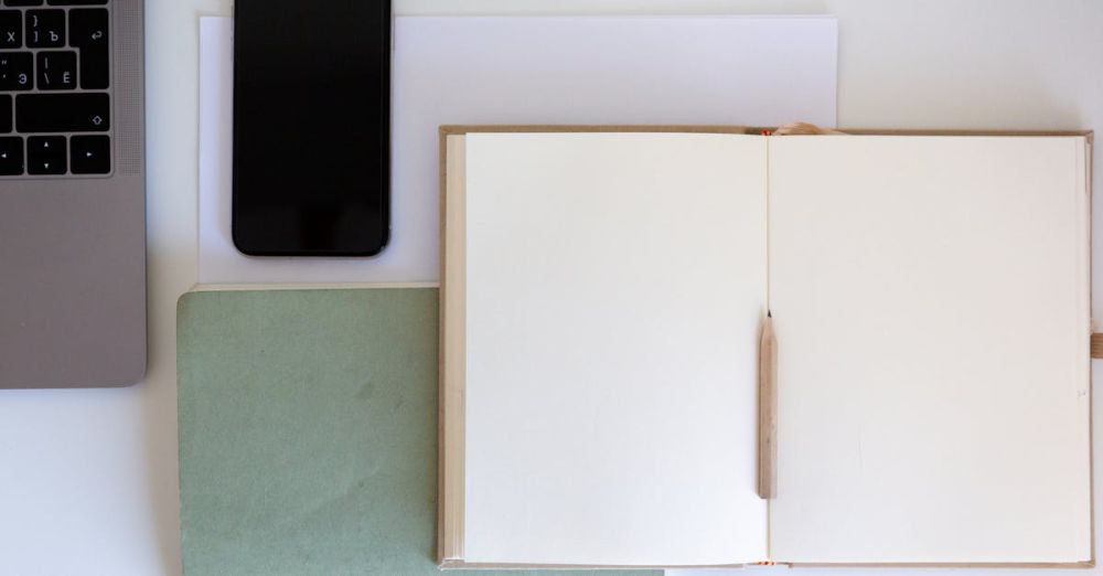 Stationery Supplies - Top view of a minimalist workspace featuring an open notebook, pencil, laptop, and smartphone on a white desk.