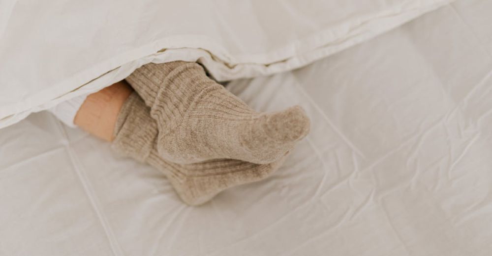 Sleep Socks - Comfortable feet in woolen socks peek out under a soft white linen blanket on a bed.