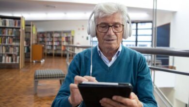 Audiobook Subscriptions - Senior man wearing headphones, using a tablet in a library setting, engaged in digital learning.