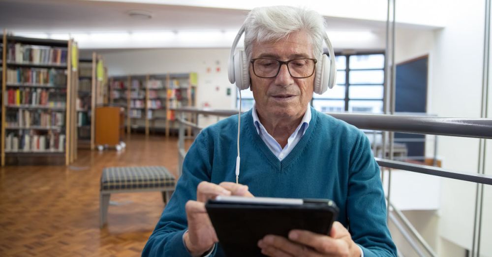 Audiobook Subscriptions - Senior man wearing headphones, using a tablet in a library setting, engaged in digital learning.