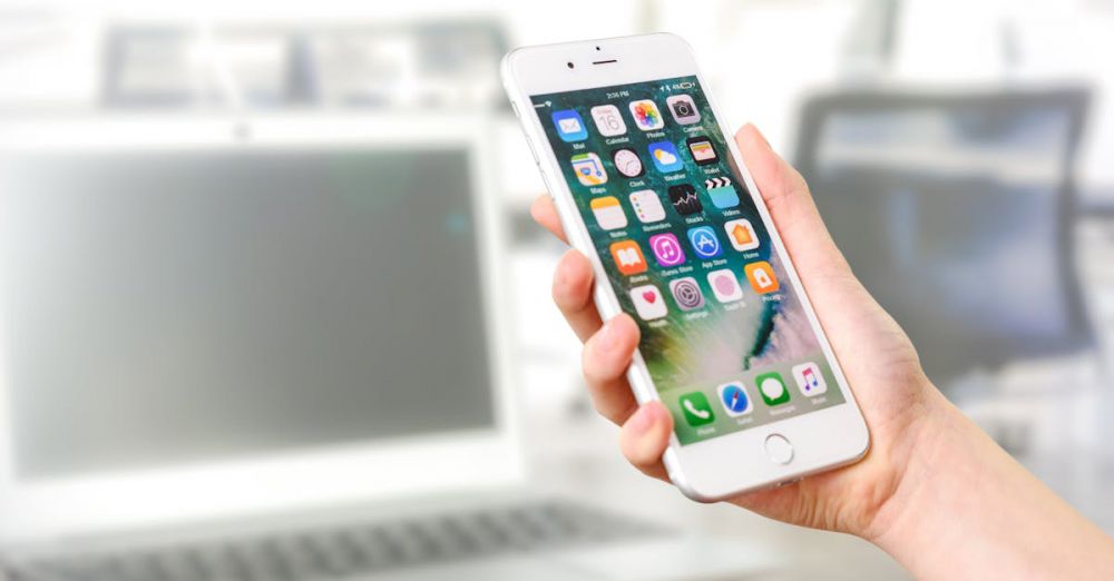 Productivity Apps - Close-up view of a smartphone displaying apps, held by a hand, with a blurred laptop in the background.