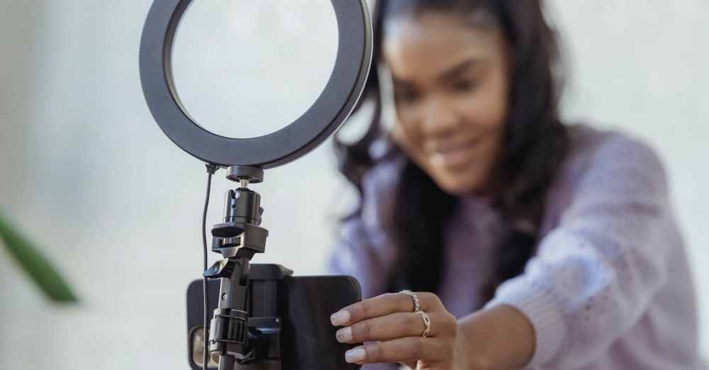 Phone Tripods - Cheerful young African American female blogger in stylish sweater smiling while setting up camera of smartphone attached to tripod with ring light before recording vlog