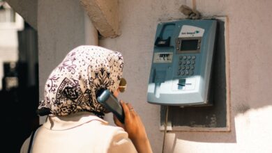 Phone Stands - A Muslim woman in hijab stands at an outdoor vintage payphone during the day.