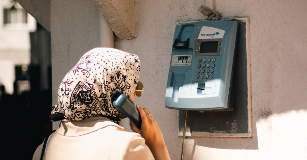 Phone Stands - A Muslim woman in hijab stands at an outdoor vintage payphone during the day.