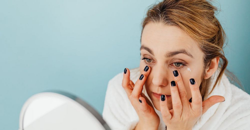 Brightening Creams - Woman applies eye cream on her under-eye area with black nails, wearing a white robe.