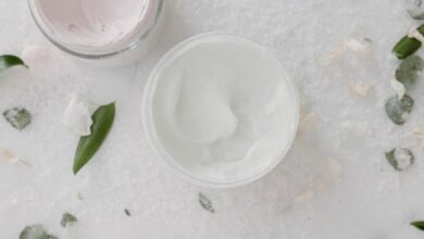 Firming Creams - Overhead shot of cosmetic cream jars surrounded by green leaves on a marble surface.