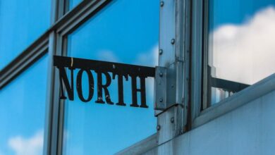 North - Reflective glass building corner with metal sign reading 'North' against blue sky and clouds.