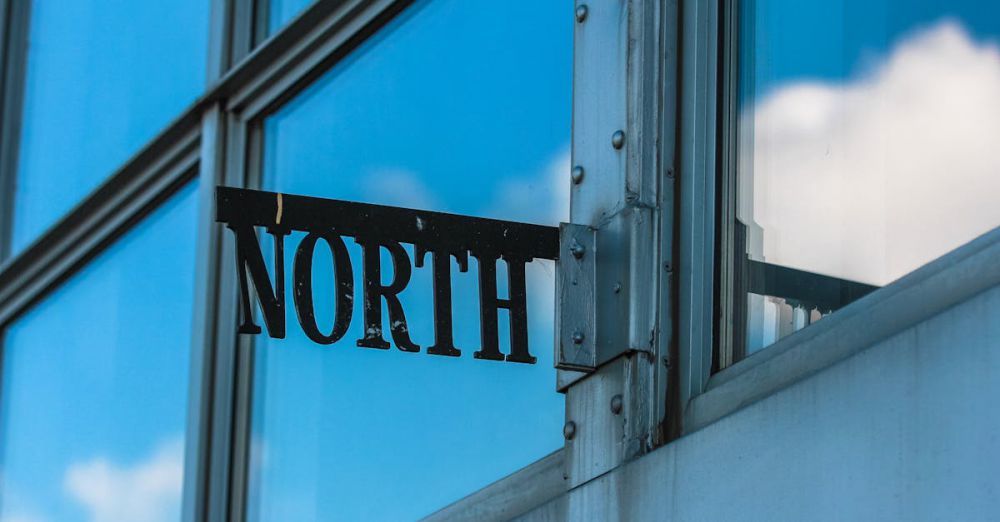 North - Reflective glass building corner with metal sign reading 'North' against blue sky and clouds.