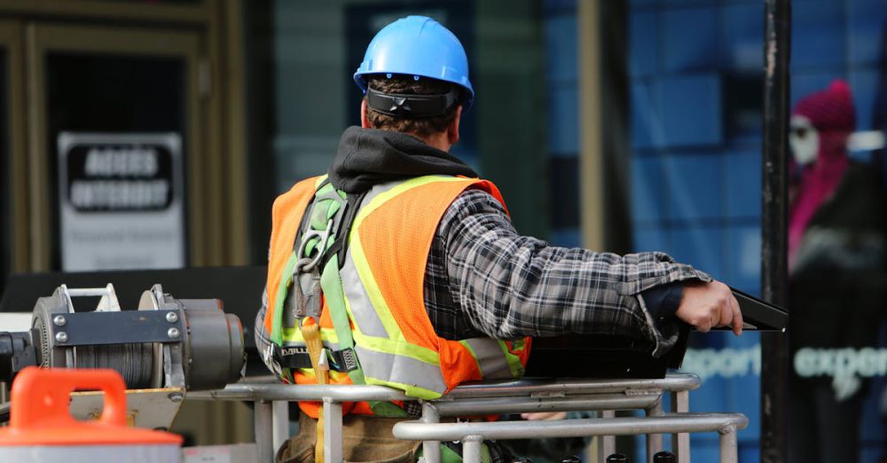 Safety - A construction worker wearing a blue helmet and safety vest operates equipment at an urban construction site.