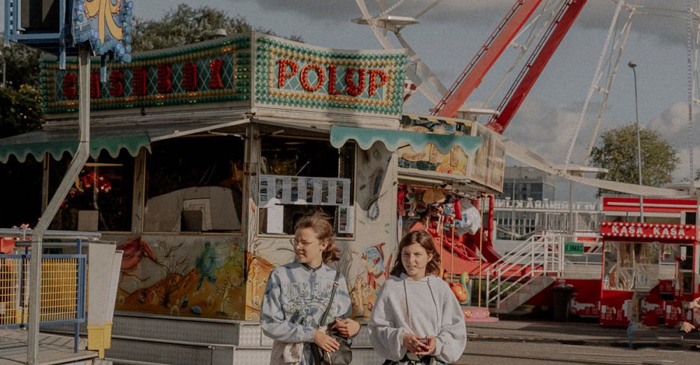 Large Groups - Two teenagers walk by colorful amusement rides at a fairground.