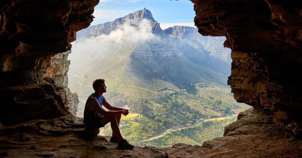 Exploration - A man sitting in a cave overlooking a majestic mountain landscape under daylight.