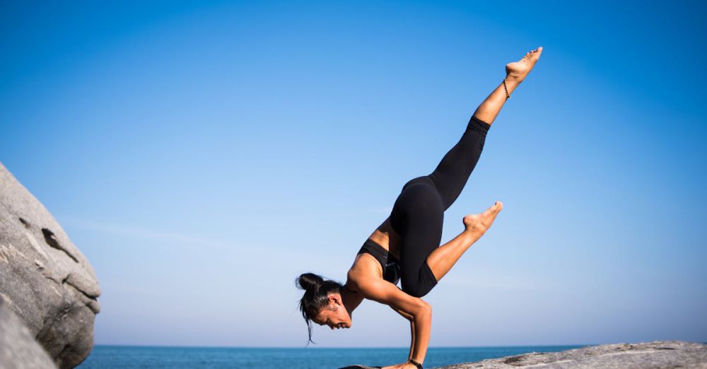 Health - Woman performing a yoga pose on a rocky beach with ocean view and clear blue sky.