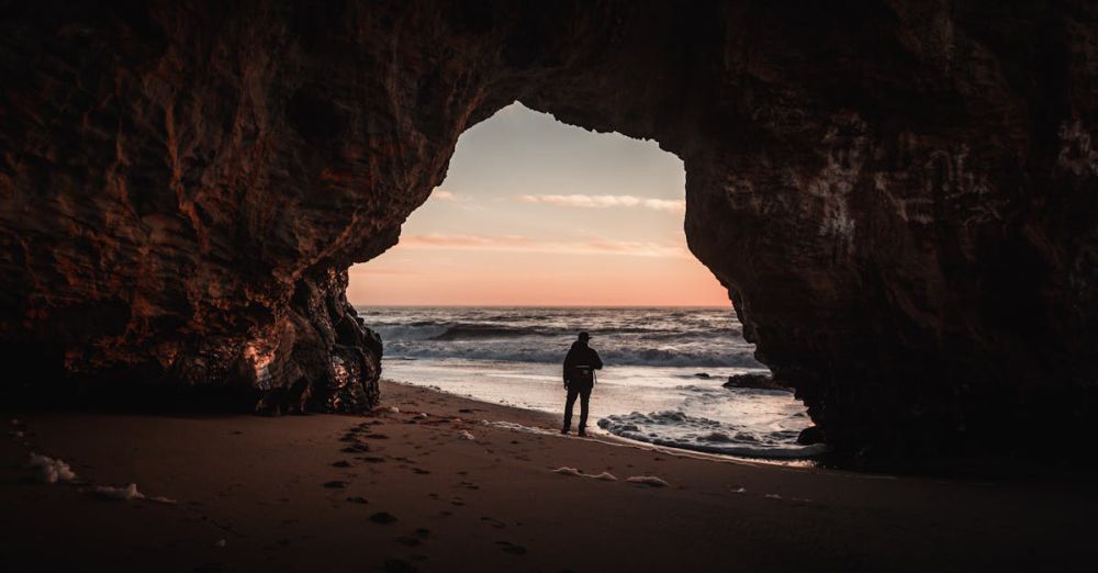 Hidden Gems - A person stands in a cave overlooking the ocean at sunset, creating a dramatic silhouette.