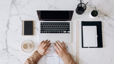 Choosing Sites - Top view of a stylish home office desk with a laptop, planner, and coffee cup, showing hands on a blueprint.