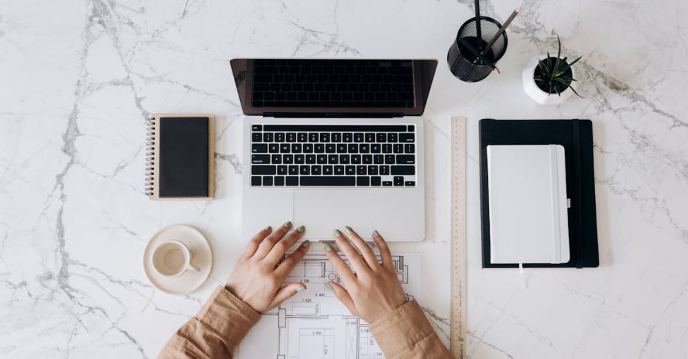 Choosing Sites - Top view of a stylish home office desk with a laptop, planner, and coffee cup, showing hands on a blueprint.