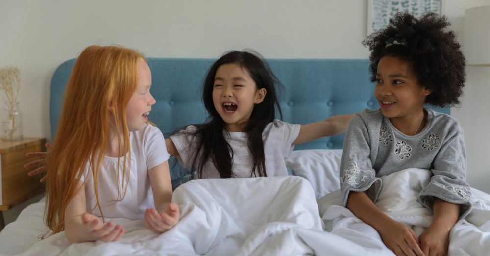 Best Time - Three happy girls bonding and laughing together in bed during a cozy sleepover.