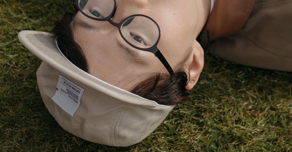 Golf Photography - A playful portrait of a young man lying on grass with a golf ball in his mouth, wearing glasses and a cap.