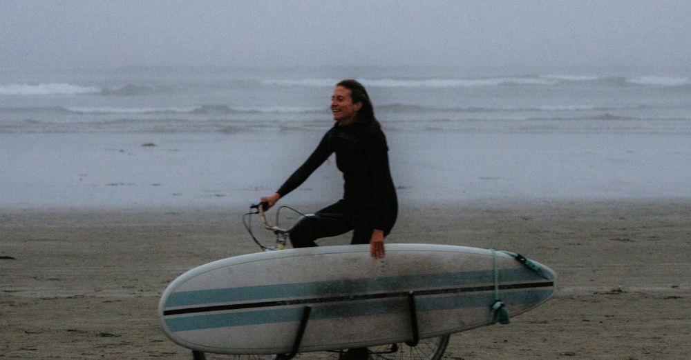 Coastal Cycling - Woman cycling along a foggy beach with a surfboard, embracing adventure and freedom.