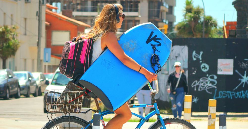 Multi-Day Cycling - A woman on a bicycle carries a surfboard and backpack through a bustling city street.