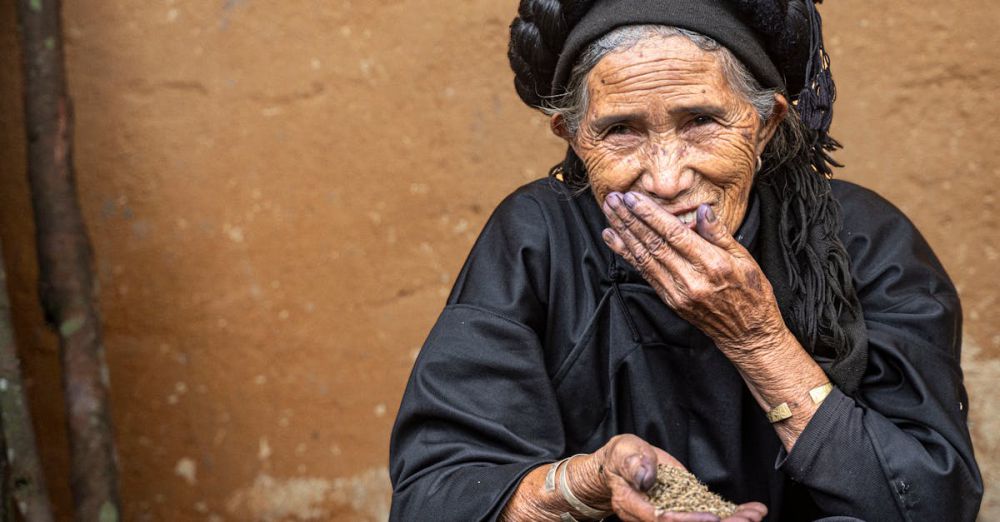 Indigenous Experiences - Smiling elderly woman in black traditional attire holding grains in Vietnam village.