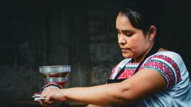 Indigenous Cuisine - A woman in traditional attire grinds coffee beans using a manual grinder in a dark setting.