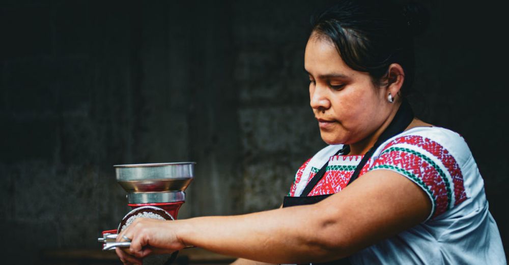 Indigenous Cuisine - A woman in traditional attire grinds coffee beans using a manual grinder in a dark setting.