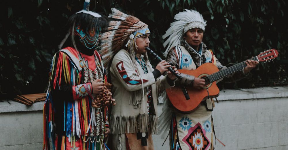 Indigenous Music - Native American musicians in traditional attire playing flute and guitar outdoors.