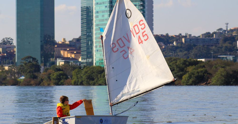 Paddling In Wind - A child sails a small boat on a city lake, framed by modern skyscrapers and lush greenery.