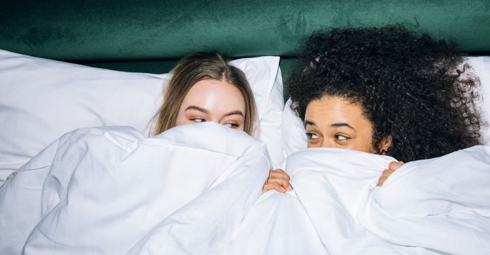 Best Time - Two young women enjoy a fun sleepover, peeking from under white bedding with smiles.