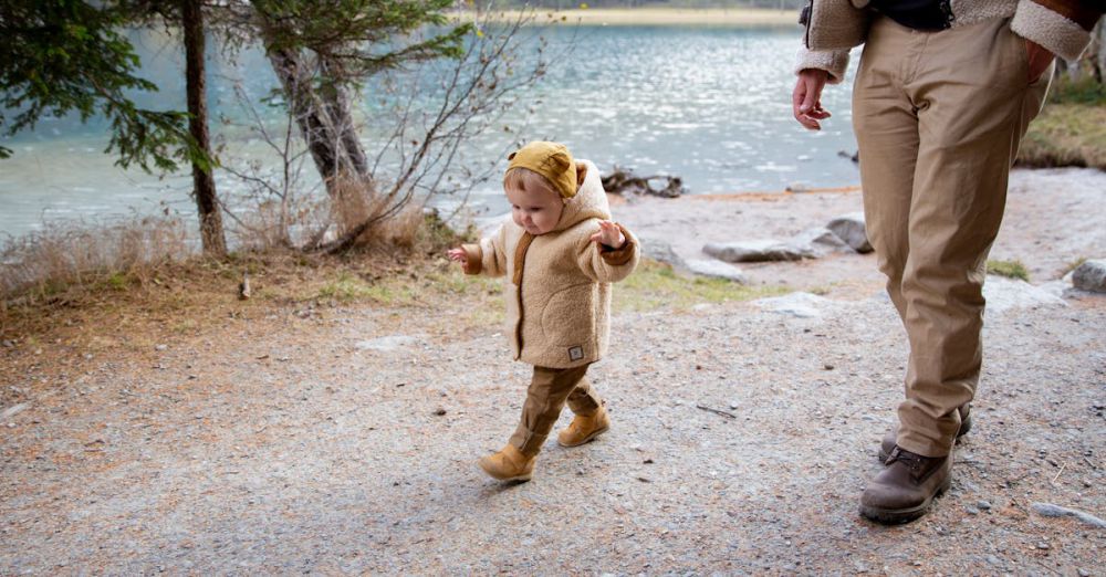 Family Trails - An adorable toddler in a jacket walks along a picturesque lakeside trail with an adult.