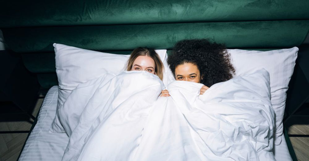 Best Time - Two young women in bed share a joyful moment, wrapped in cozy white bedding indoors.