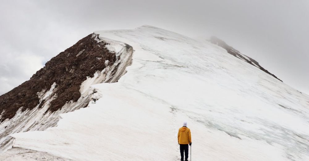 Solo Climbers - A lone climber in winter gear ascends a snowy mountain peak under cloudy skies.