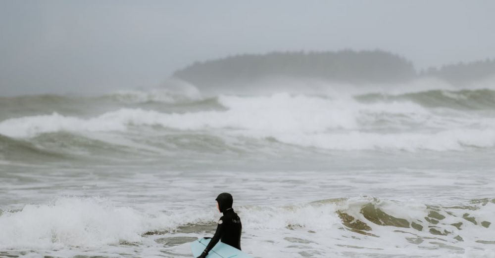 Cold Water Surfing - A lone surfer with a surfboard stands on a misty beach, preparing to ride the ocean waves.