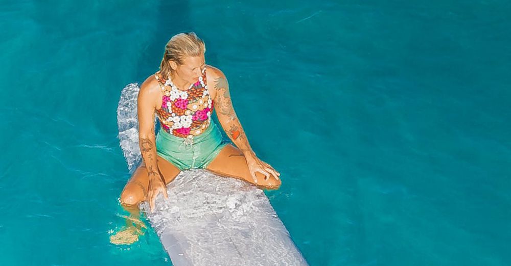 Solo Surfing - An adult female surfer in a floral top sits on a surfboard in Hawaii's clear blue ocean.