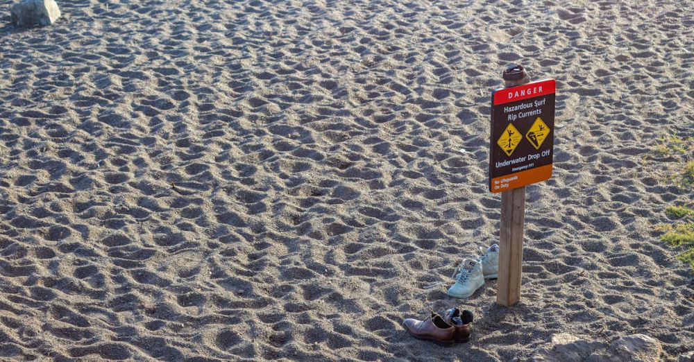 Rip Currents - A beach warning sign stands on the sandy shore with two pairs of shoes nearby, emphasizing caution.