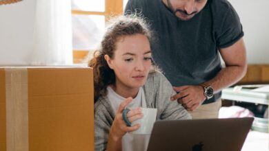 New Brunswick Surfing - Thoughtful woman surfing internet on netbook while sitting with cup of hot drink near ethnic boyfriend in watch and casual wear standing near cardboard boxes after moving into new flat