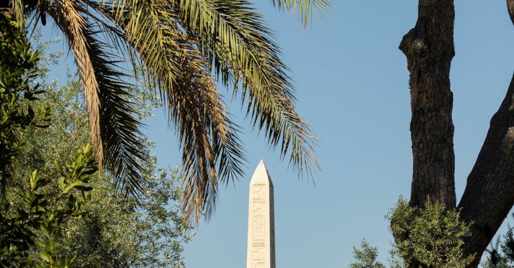 Scenic Locations - A historic obelisk surrounded by palm trees and greenery in Rome, under a clear blue sky.