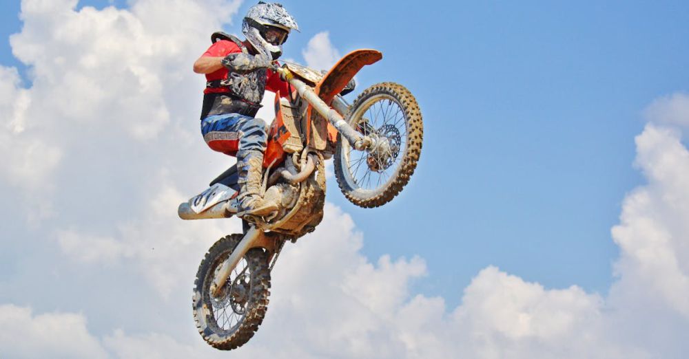 Extreme Sports - Motocross rider performs an airborne jump under a bright blue sky with clouds.