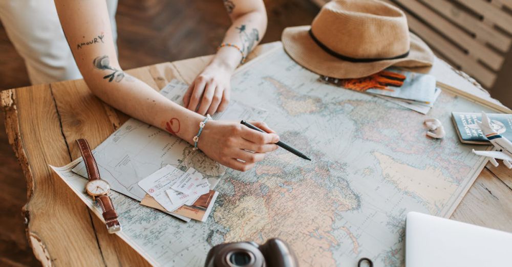 Underrated Routes - Tattooed woman planning a trip with a world map, passports, and travel essentials on a wooden table.