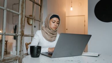 Experiences - Muslim woman in hijab typing on a laptop indoors with a coffee mug nearby at a table.