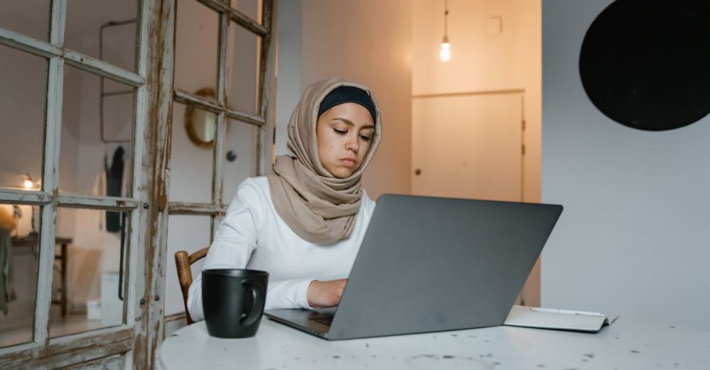 Experiences - Muslim woman in hijab typing on a laptop indoors with a coffee mug nearby at a table.